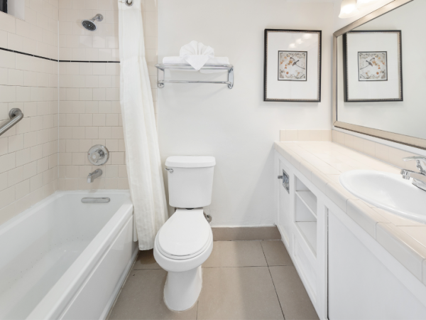 A compact bathroom with a white bathtub/shower, toilet, and long vanity mirror, two framed pictures, and a clean, gray-tiled floor.