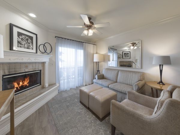 Cozy living room with a fireplace, light gray sofa, matching armchairs, ottomans, and a large wall mirror; soft lighting and sheer curtains.