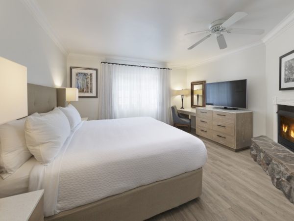 A bright hotel-style bedroom with a king bed, white bedding, a dresser, a wall-mounted TV, a fireplace, and a window with sheer curtains.
