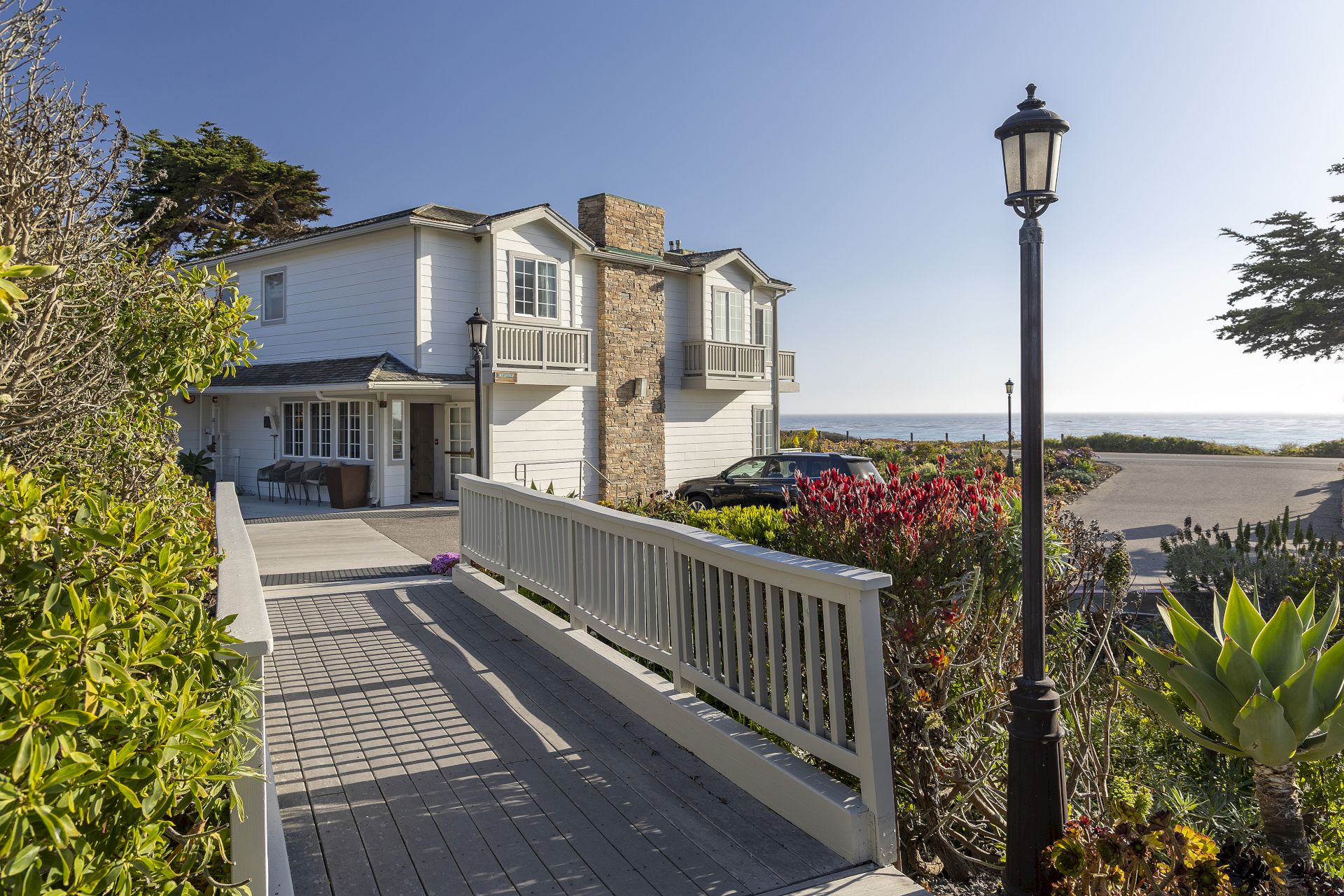Sunlit coastal home patio with white railings, flowering gardens, a black lantern post, and the ocean beyond.