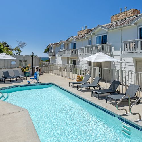 A sunny apartment complex with a rectangular pool, lounge chairs, umbrellas, and balconies overlooking the pool area. Indeed.