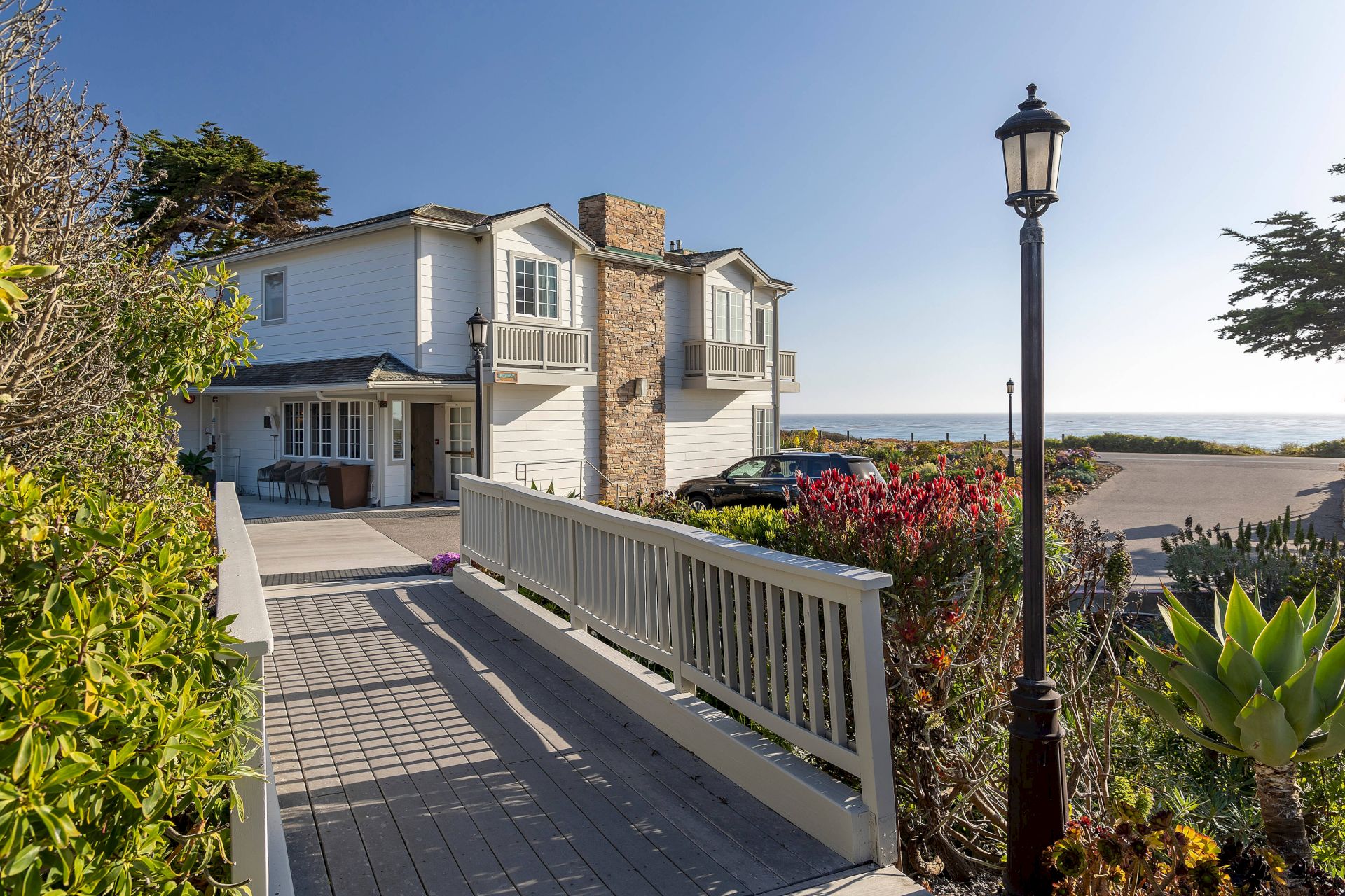 A white coastal house with balconies, garden plants, and a lantern-lit walkway beside the ocean under a clear sky.
