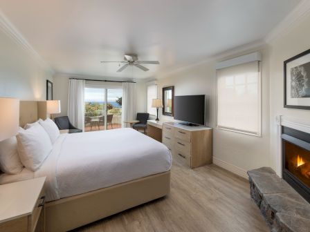 A bright hotel room with a king bed, nightstands, a dresser with a TV, a sitting area by a balcony, and a cozy fireplace in the corner.