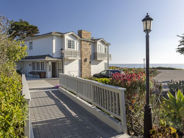 Cozy coastal house with a wooden walkway leading to a white two-story home, stone chimney, fence, lamp post, and nearby greenery by the beach.