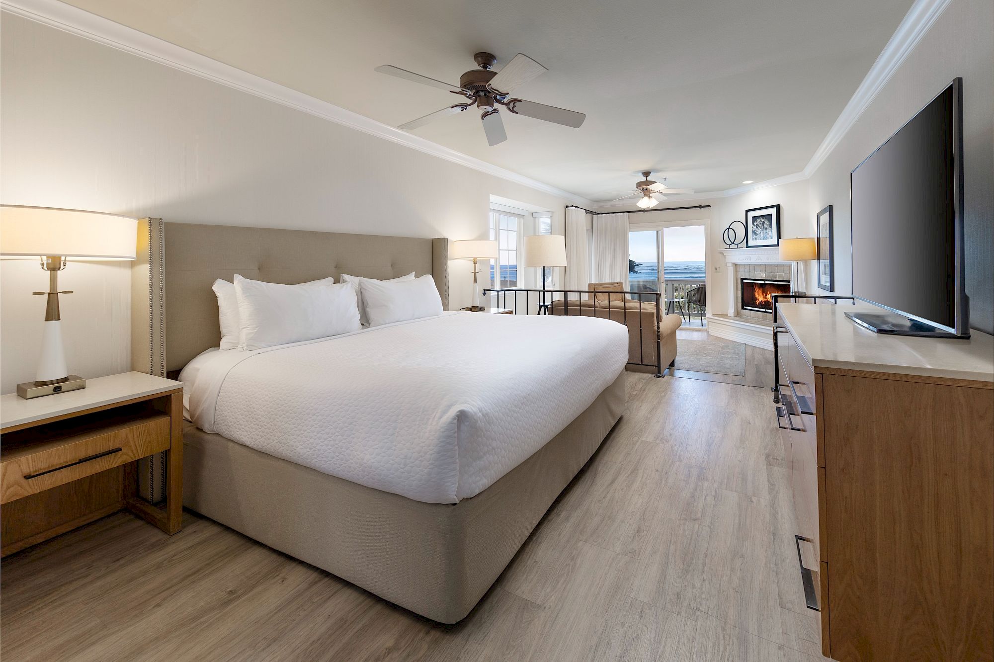 A bright hotel room with a large white bed, nightstands, a flat-screen TV, desk, seating area, ceiling fans, and light wood flooring.