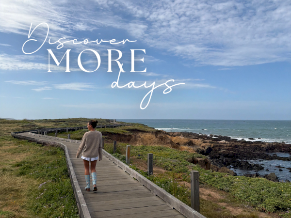 A person strolls along a wooden coastal boardwalk beside rocky shore and ocean under a cloudy blue sky, with &ldquo;Discover MORE days&rdquo; text.